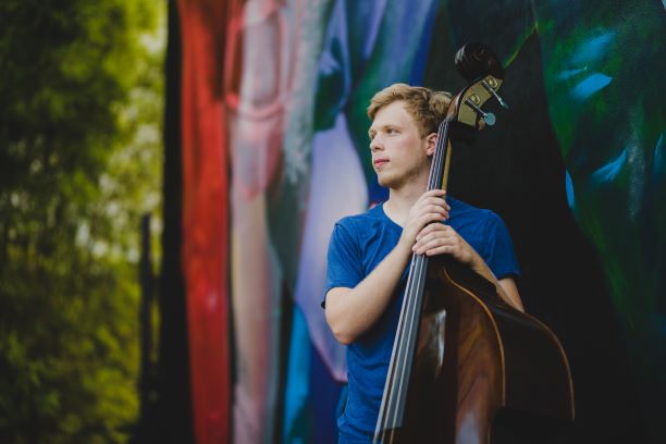 Andrew Binder holding an upright bass and looking off to the left in front of a colorful, blurred background