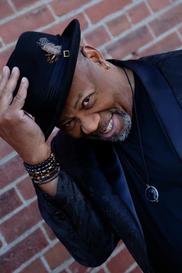 Johnnie Owens, dressed in a black suit over a black shirt, tipping a black fedora and smiling in front of a red brick wall.