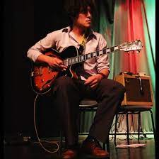 Jose Gobbo sits on a stage with a guitar and an amp.