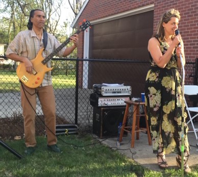 Liz Fitz Band performing outdoors. Liz is dressed in a long, floral maxi dress and is singing into a microphone. A man plays guitar behind her.