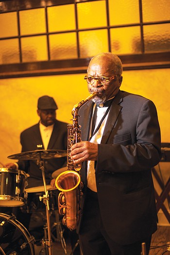 A color photograph of Virgil Rhodes dressed in a dark suit playing the saxophone on stage. A drummer is scene in the background.