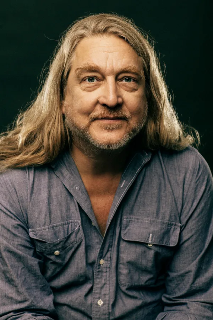 A color headshot of Tom Irwin, a man with a short beard and long, blond hair. He wears a chambray shirt.
