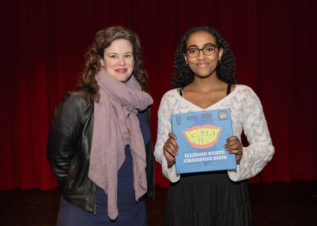 A color photo of Angelique Grandone and Yohanna Endashaw posing in front of a red stage curtain. Yohanna is holding her State Champion award.