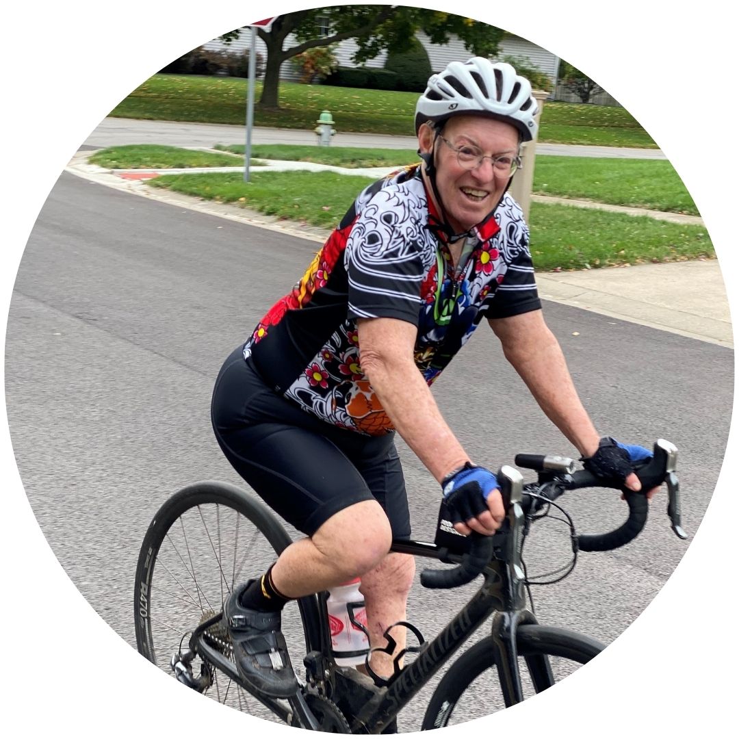 A color photo of Steve Rambach smiling for the camera as he rides a racing bicycle. He is wearing a white helmet, glasses, a black and white graphic shirt with colorful details.