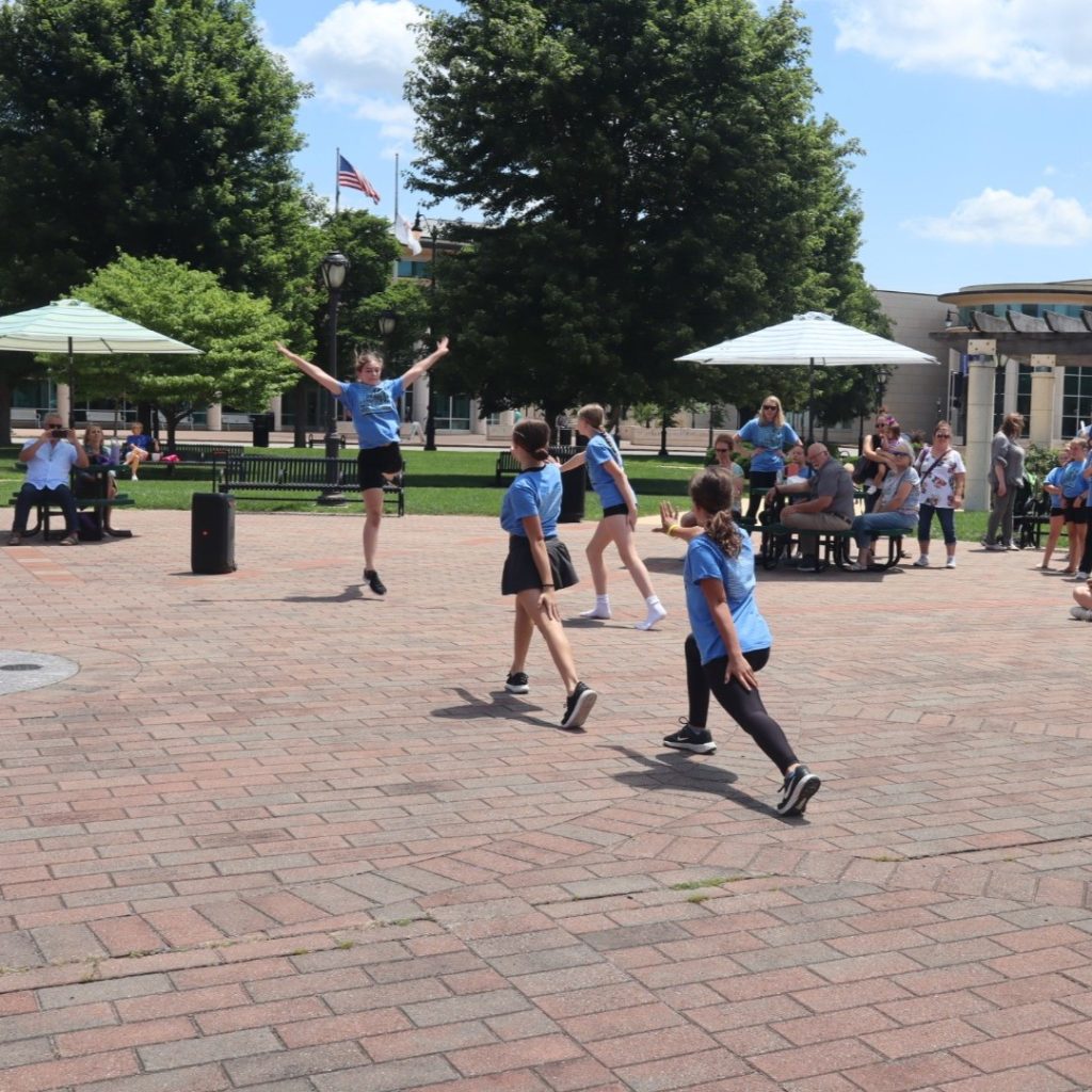 group of kids dancing on cobblestone in a park surround trees with an audience behind them sitting at tables