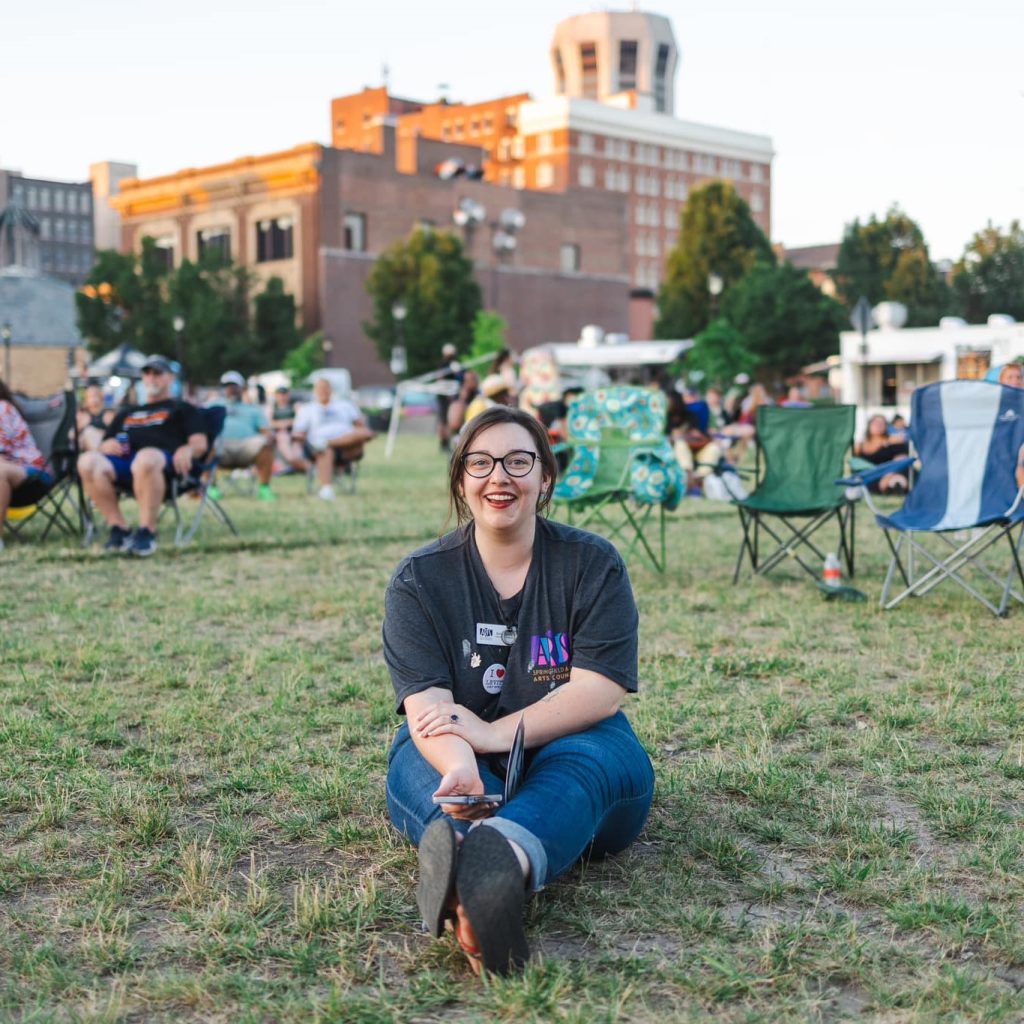 woman laughing while sitting on the grass in a park with a phone in her hands with people sitting in lawn chairs behind her and large buildings in the background.