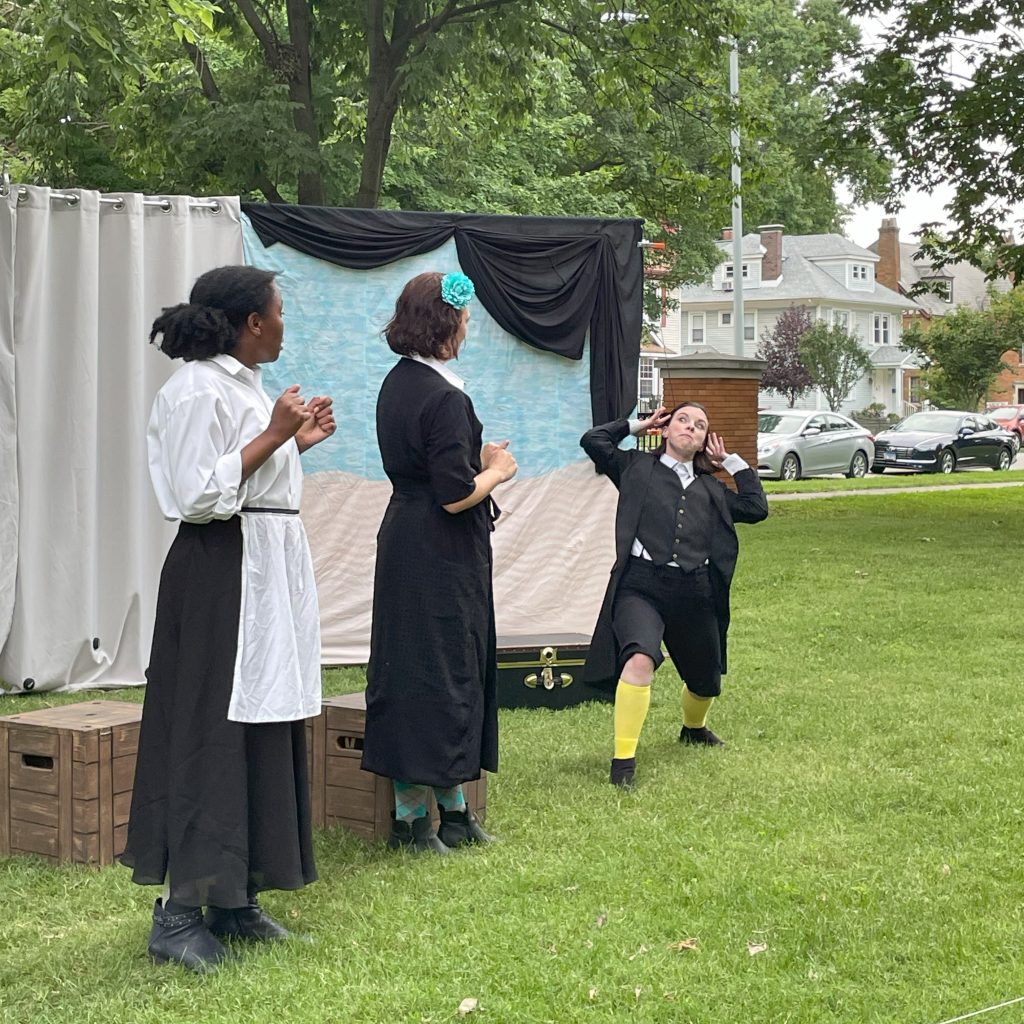 three actors performing a play in a park with a pipe and drape backdrop