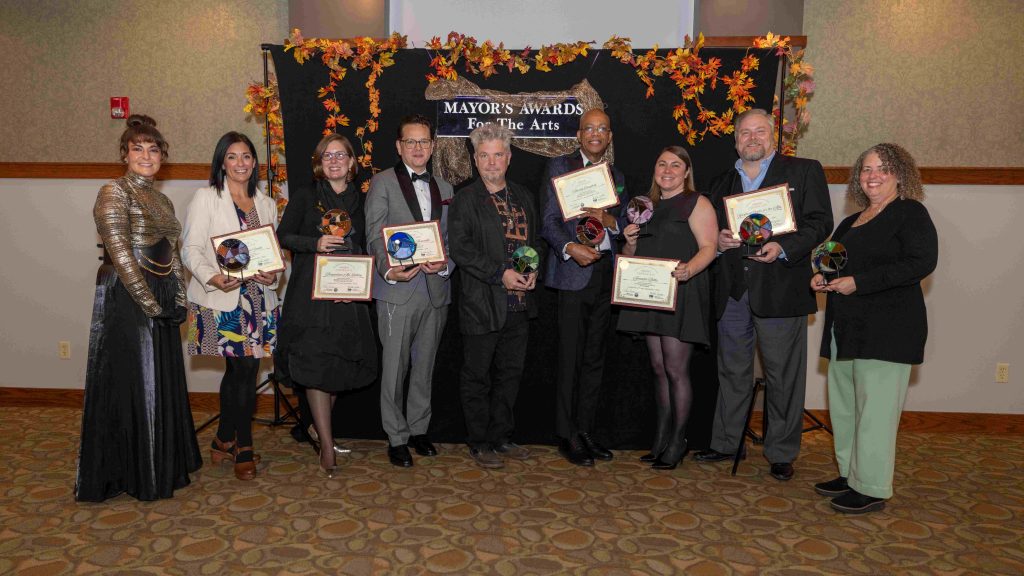 A group of people posing with awards in front of a black backdrop with fall leaves hanging down.