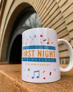 A white, ceramic mug with the 38th annual First Night Springfield logo, with colorful confetti and music notes sitting on a concrete ledge in front of an archway.