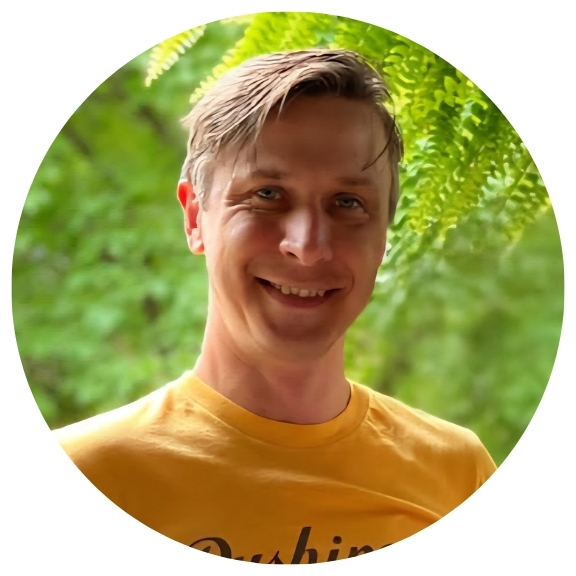 Man in yellow t-shirt smiling while standing in front of green foliage.