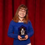 Young woman wearing glasses, in a blue sweater, holing an award.
