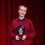 Young woman with short hair wearing a red sweater and holding an award.